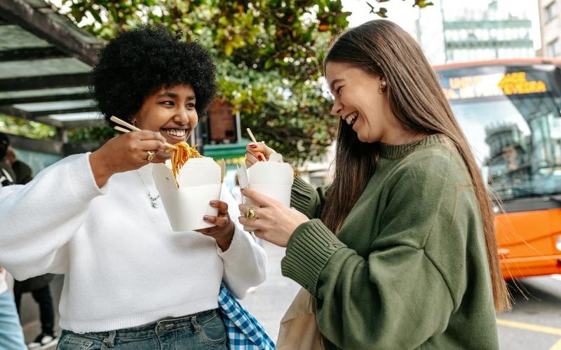 Two women laughing and eating street food in the city.